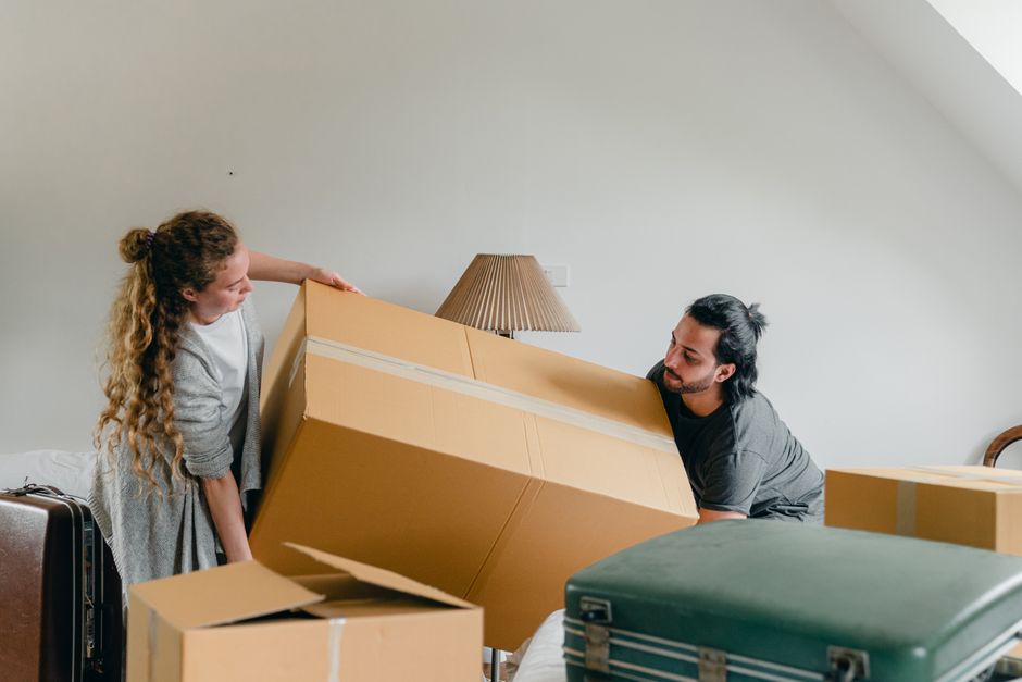 An interior scene showing a man and woman preparing for a home relocation within a residential property. The woman, with curly red hair, holds a pink and white striped piece of clothing or fabric, sta